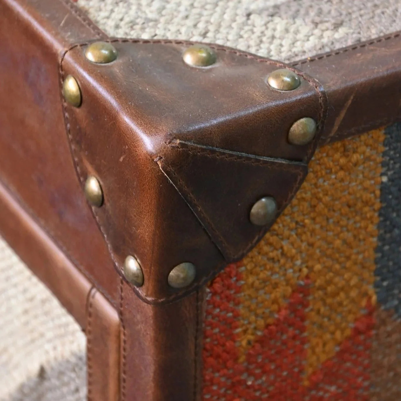 Close-up of a brown leather suitcase corner with brass studs on a textured fabric background.